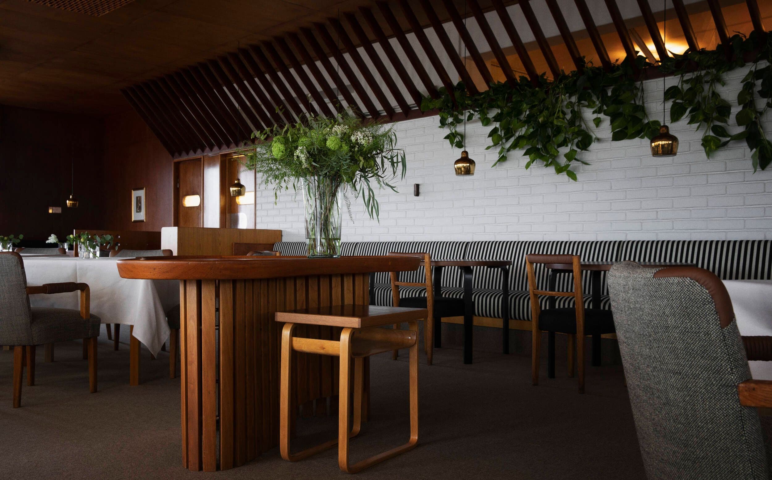 The main dining hall is framed by a long sofa. Hung in a row, the original Golden Bell pendants bring a beautiful glow on the bespoke Artek tables. The free-form serving table ‘Alvar’s Table’ sits in the middle of the room.